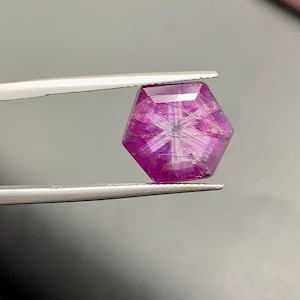 以下が含まれることがあります： ピンセットで挟まれた、ファセット加工された六角形の宝石。宝石は深いマゼンタ色で、内部に星のような模様が見えます。宝石のファセットが光を捉え、きらめく効果を生み出しています。背景はぼやけたグレーです。