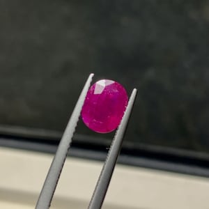 May include: A vibrant, oval-cut ruby gemstone with a deep magenta-pink hue and faceted surface, held securely by a pair of silver-toned metal tweezers. The gemstone displays internal clarity and light reflection across its polished, multi-faceted top.