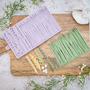 Puede incluir: Un molde de silicona morado con varios diseños de hojas y hierba. El molde está sobre una tabla de cortar de madera con hojas y hierba de fondant verde y amarillo hechas con el molde.