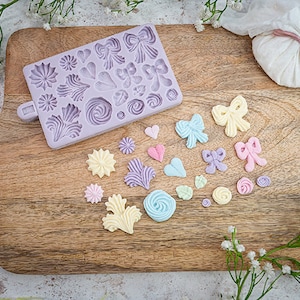 May include: A purple silicone mold with various designs for making decorative sugar paste flowers and bows. The mold is on a wooden cutting board with several finished sugar paste flowers and bows in pastel colors.