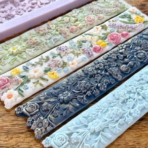 May include: Close-up of several floral-patterned moulds in various colours, including light green, white, navy blue, and light blue. The moulds feature detailed designs of roses, daisies, and other flowers, ideal for crafting projects.