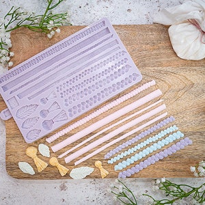 May include: A purple silicone mould with various designs for creating cake decorations. The mould includes designs for leaves, tassels, beads, and different types of borders. There are also several finished decorations made with the mould, including pink and blue beaded chains, pink and white tassels, and yellow tassels.