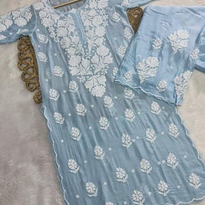 Puede incluir: Un conjunto bordado de dos piezas en azul claro. La parte superior de manga larga y los pantalones a juego presentan un intrincado bordado floral blanco. La parte superior tiene un dobladillo festoneado y se muestra en una percha de madera.