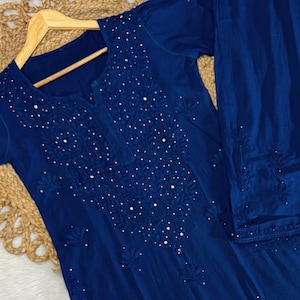 Op de afbeelding: Een marineblauw, geborduurd tweedelig outfit. De top heeft een V-hals en is versierd met kleine witte versieringen en bloemenborduurwerk. Een bijpassende broek is inbegrepen. Een kleine, ronde borduurring met bloemenontwerpen staat op de voorgrond.