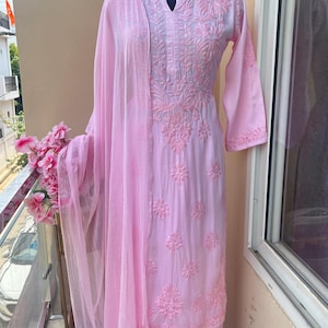 May include: A light pink embroidered salwar kameez set displayed on a mannequin. The set includes a long-sleeved tunic, matching pants, and a sheer scarf. The tunic and pants feature detailed floral embroidery. The outfit is displayed on a balcony.