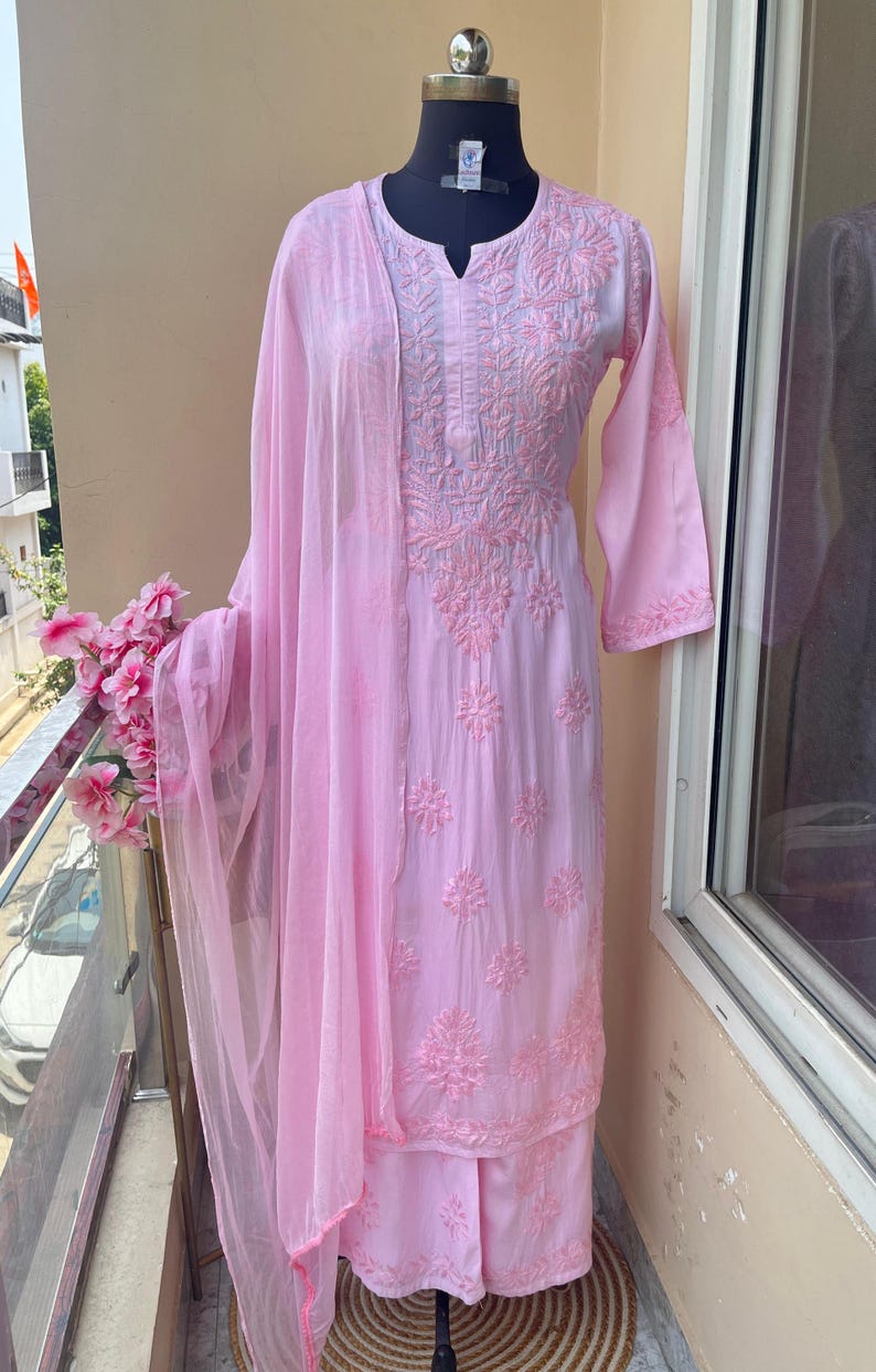 May include: A light pink embroidered salwar kameez set displayed on a mannequin. The set includes a long-sleeved tunic, matching pants, and a sheer scarf. The tunic and pants feature detailed floral embroidery. The outfit is displayed on a balcony.