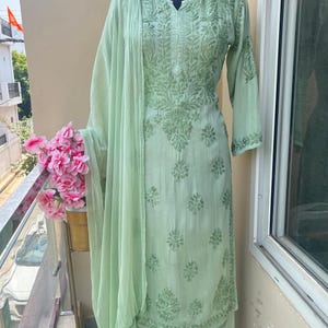 May include: A light green embroidered salwar kameez set, including a tunic, pants, and a matching scarf. The fabric features detailed floral patterns. The outfit is displayed on a mannequin.