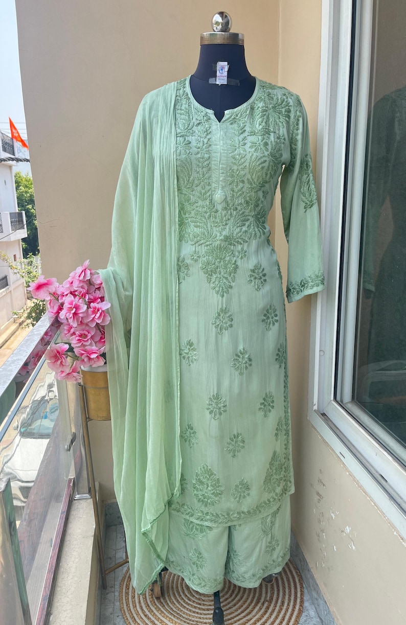 May include: A light green embroidered salwar kameez set, including a tunic, pants, and a matching scarf. The fabric features detailed floral patterns. The outfit is displayed on a mannequin.