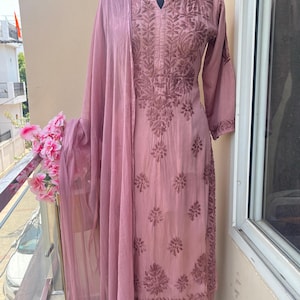 May include: A dusty rose-colored salwar kameez set displayed on a mannequin. The ensemble features intricate floral embroidery on the tunic, pants, and matching sheer dupatta. The outfit is made of a soft fabric and is suitable for special occasions.