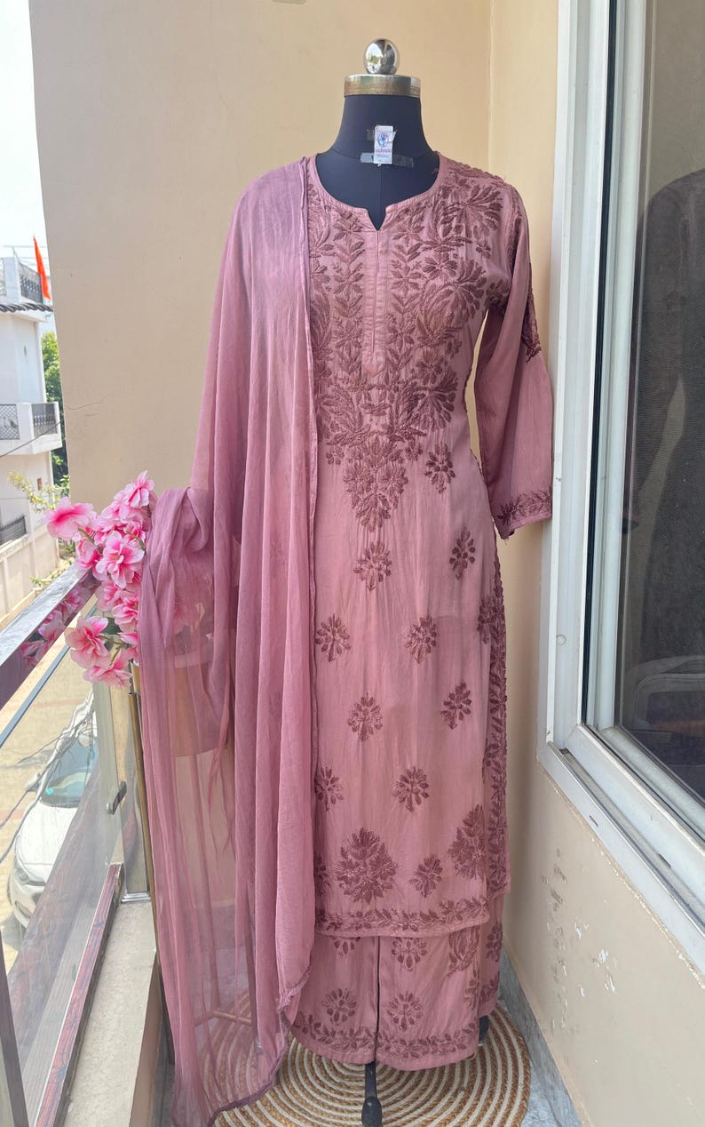 May include: A dusty rose-colored salwar kameez set displayed on a mannequin. The ensemble features intricate floral embroidery on the tunic, pants, and matching sheer dupatta. The outfit is made of a soft fabric and is suitable for special occasions.