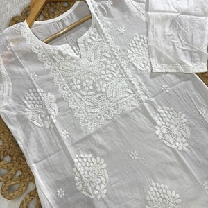 May include: A white, long-sleeved tunic with intricate floral embroidery. The tunic features a round neckline with a small slit and is displayed on a wooden hanger. The embroidery is concentrated around the neckline, chest, and sleeves.