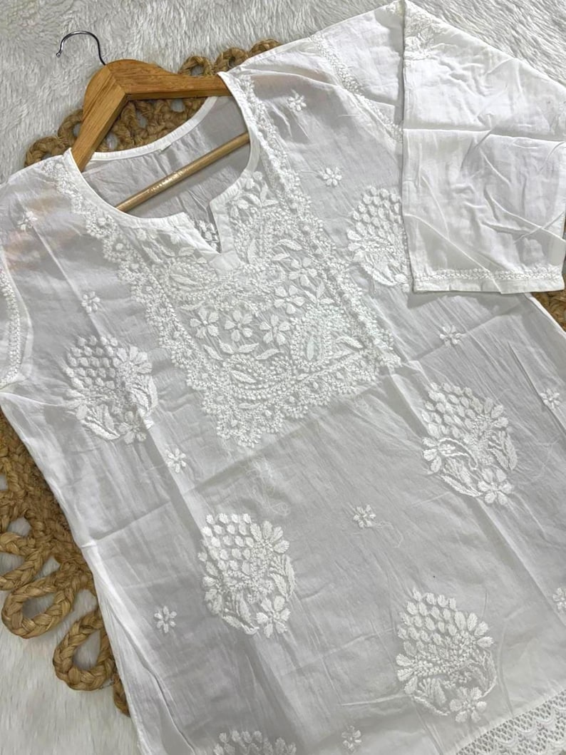 May include: A white, long-sleeved tunic with intricate floral embroidery. The tunic features a round neckline with a small slit and is displayed on a wooden hanger. The embroidery is concentrated around the neckline, chest, and sleeves.