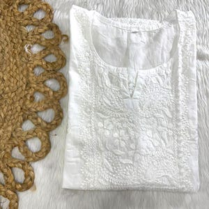 May include: A white, embroidered tunic with a round neckline and a small V-shaped opening. The tunic features detailed floral embroidery across the front and around the neckline. The garment is folded and placed on a white, fluffy surface, with a woven jute placemat on the left.