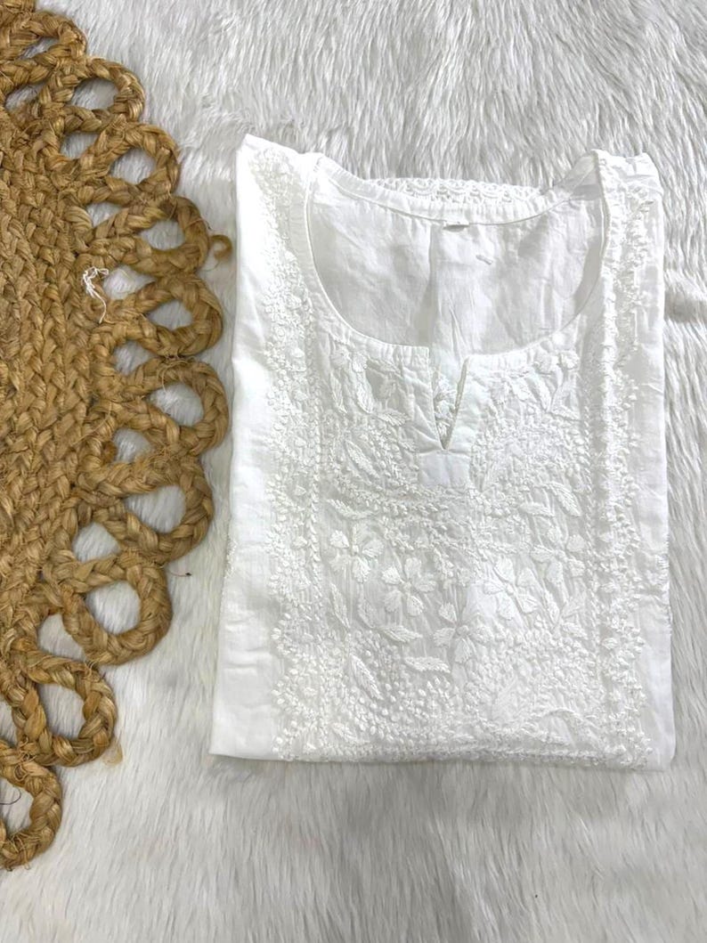 May include: A white, embroidered tunic with a round neckline and a small V-shaped opening. The tunic features detailed floral embroidery across the front and around the neckline. The garment is folded and placed on a white, fluffy surface, with a woven jute placemat on the left.