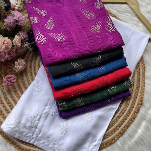 May include: A stack of six embroidered tunics in various colors, including purple, black, blue, red, green, and purple. Each tunic features a paisley design with rhinestones. The tunics are folded and stacked on top of a white tunic with a floral design.