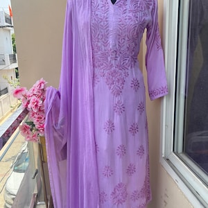 May include: A light purple embroidered salwar kameez set, displayed on a mannequin. The set includes a long-sleeved tunic, matching pants, and a sheer scarf. The intricate floral embroidery covers the tunic and pants. The outfit is displayed on a balcony.