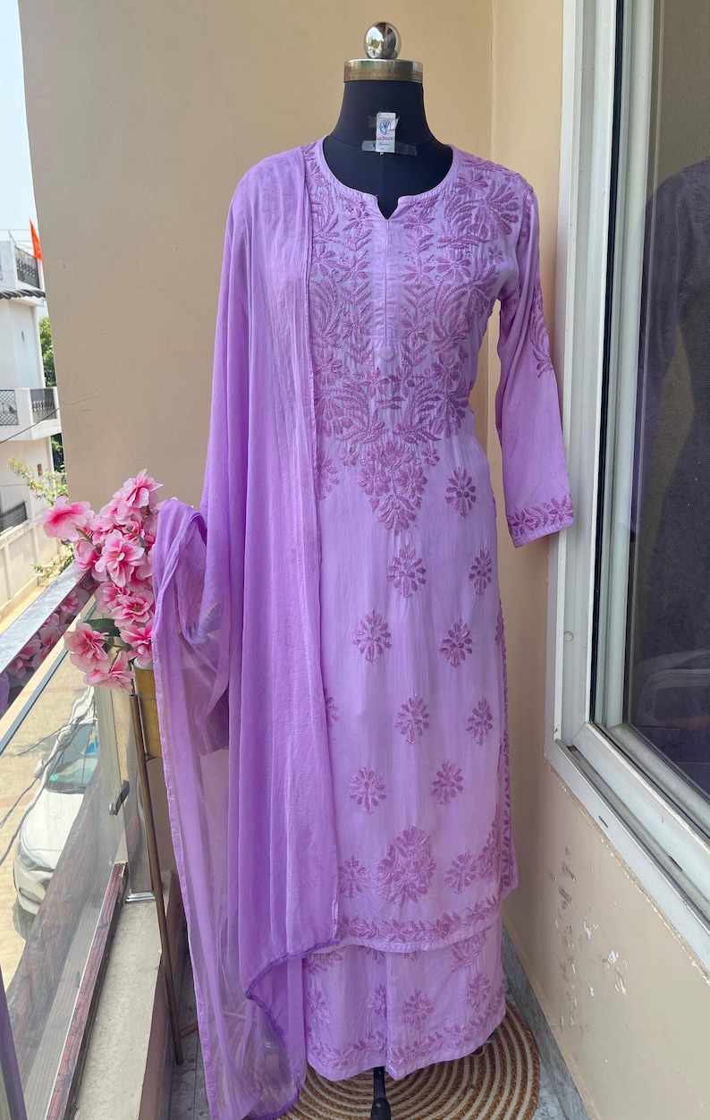 May include: A light purple embroidered salwar kameez set, displayed on a mannequin. The set includes a long-sleeved tunic, matching pants, and a sheer scarf. The intricate floral embroidery covers the tunic and pants. The outfit is displayed on a balcony.