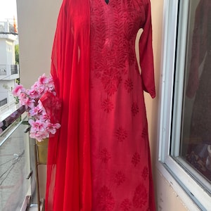 May include: A vibrant red embroidered salwar kameez ensemble. The outfit includes a long-sleeved tunic, matching pants, and a sheer scarf. The fabric is adorned with detailed floral patterns. The outfit is displayed on a mannequin.