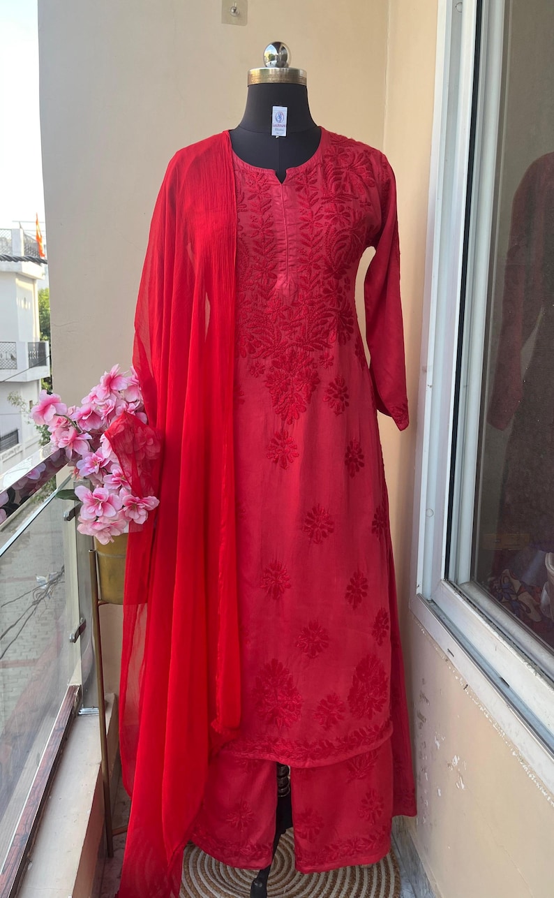 May include: A vibrant red embroidered salwar kameez ensemble. The outfit includes a long-sleeved tunic, matching pants, and a sheer scarf. The fabric is adorned with detailed floral patterns. The outfit is displayed on a mannequin.