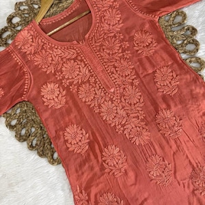 以下が含まれることがあります： サンゴ色のチュニックで、繊細な花の刺繍が施されています。長袖のトップスは丸いネックラインで、胸と袖に詳細な模様があります。木製のハンガーにかけられており、繊細な生地とデザインが際立っています。