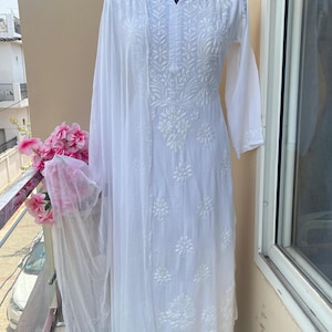 May include: A white embroidered kurta suit displayed on a mannequin. The suit includes a long-sleeved tunic, palazzo pants, and a sheer scarf. The fabric is adorned with detailed floral embroidery. The outfit is displayed on a balcony with pink flowers.