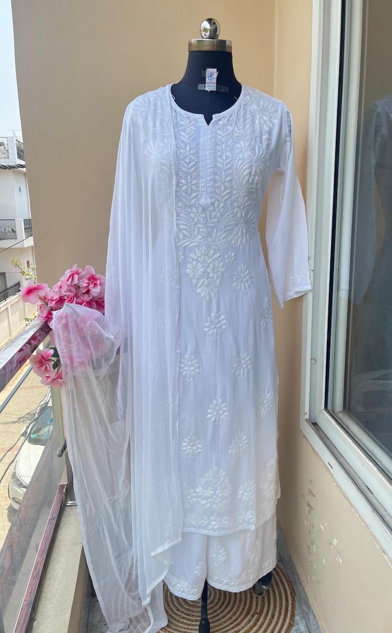 May include: A white embroidered kurta suit displayed on a mannequin. The suit includes a long-sleeved tunic, palazzo pants, and a sheer scarf. The fabric is adorned with detailed floral embroidery. The outfit is displayed on a balcony with pink flowers.