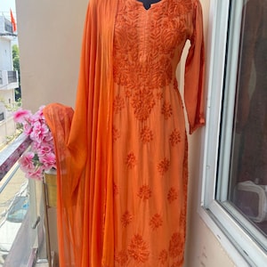 May include: An orange embroidered salwar kameez ensemble displayed on a mannequin. The outfit includes a long-sleeved tunic, matching pants, and a draped scarf. The fabric features detailed floral embroidery.