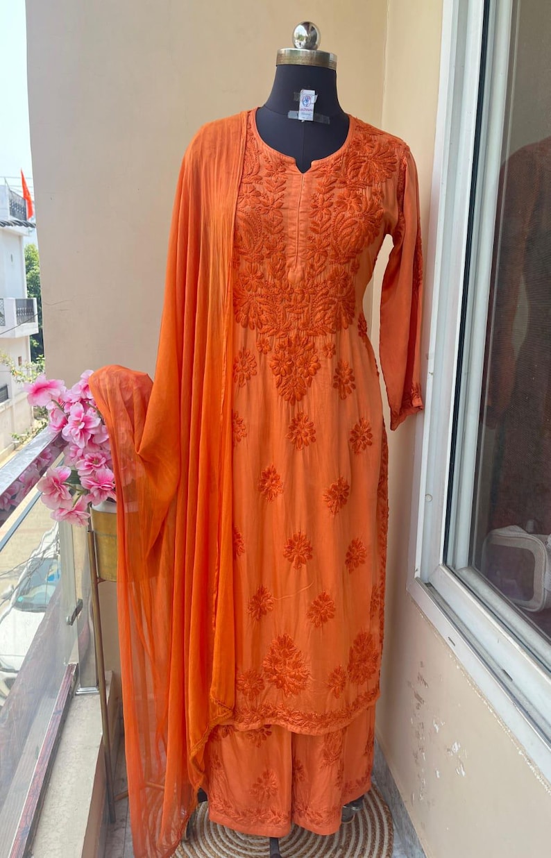 May include: An orange embroidered salwar kameez ensemble displayed on a mannequin. The outfit includes a long-sleeved tunic, matching pants, and a draped scarf. The fabric features detailed floral embroidery.