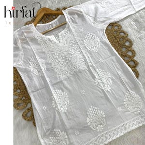 Puede incluir: Túnica blanca con intrincado bordado floral. La parte superior de manga larga presenta patrones detallados alrededor del escote, las mangas y el dobladillo. La palabra "hirfat" es visible en la esquina superior izquierda.