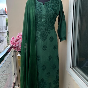 May include: Emerald green embroidered salwar kameez set displayed on a mannequin. The set includes a long-sleeved tunic, matching palazzo pants, and a sheer scarf. The fabric features detailed floral embroidery. The outfit is displayed on a balcony.