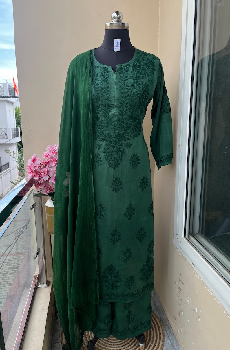 May include: Emerald green embroidered salwar kameez set displayed on a mannequin. The set includes a long-sleeved tunic, matching palazzo pants, and a sheer scarf. The fabric features detailed floral embroidery. The outfit is displayed on a balcony.