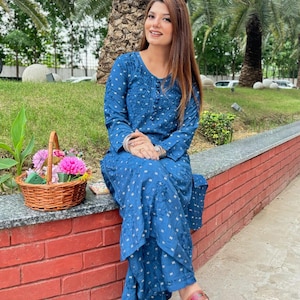 May include: A woman wearing a blue patterned long-sleeved top and matching trousers. She is sitting on a brick wall with a wicker basket of flowers beside her.