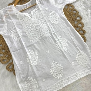 May include: White tunic with intricate floral embroidery. The long-sleeved top features a notched neckline and delicate lace trim along the hemline. The embroidery is detailed and covers the front and sleeves.