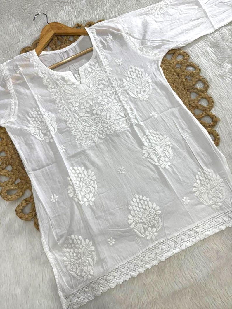 May include: White tunic with intricate floral embroidery. The long-sleeved top features a notched neckline and delicate lace trim along the hemline. The embroidery is detailed and covers the front and sleeves.