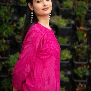 May include: A woman wearing a pink embroidered kurta with long sleeves and a round neckline. She is wearing pearl drop earrings and has long dark hair.