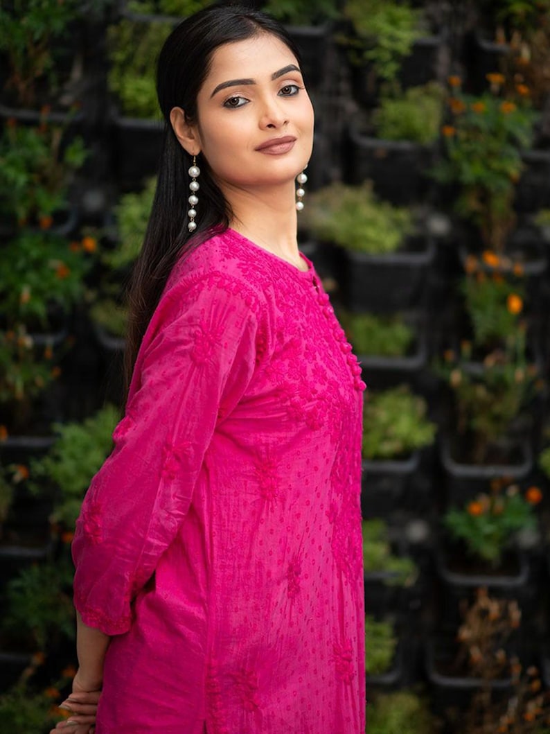 May include: A woman wearing a pink embroidered kurta with long sleeves and a round neckline. She is wearing pearl drop earrings and has long dark hair.