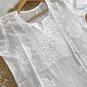 May include: White, embroidered tunic with short sleeves. The tunic features intricate floral and leaf patterns around the neckline and on the front. The garment is hanging on a wooden hanger against a neutral background.