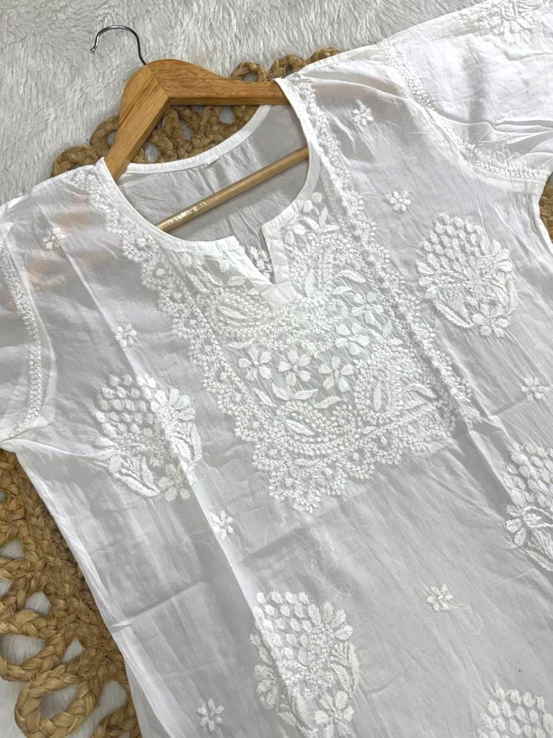 May include: White, embroidered tunic with short sleeves. The tunic features intricate floral and leaf patterns around the neckline and on the front. The garment is hanging on a wooden hanger against a neutral background.