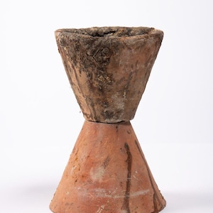 May include: A two-part terracotta pot with a conical base and a flared, bowl-shaped top. The top section has a rough, weathered texture, while the base is smooth with dark streaks. The pot is likely for decorative or functional use.