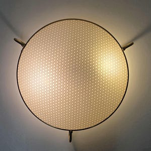 Könnte beinhalten: Eine runde, cremefarbene Leuchte mit einem gewebten Muster. Sie wird von einem dunklen Metallring umrahmt und hat drei gebogene Metallhalterungen. Das Licht ist eingeschaltet und strahlt einen warmen Schein aus.