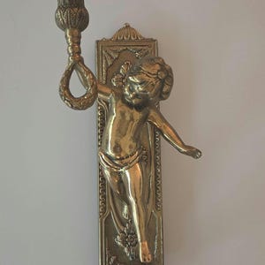 Puede incluir: Un aplique de pared de latón con una figura de querubín sosteniendo un candelabro. El aplique tiene un diseño ornamentado con detalles florales y de volutas. El querubín está en una pose dinámica, añadiendo un toque de elegancia clásica a la decoración.