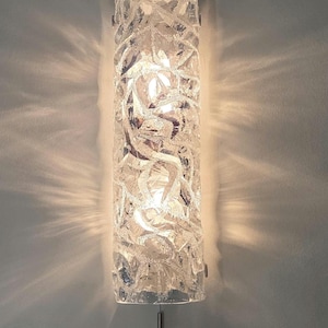 May include: A clear, textured glass wall sconce with a cylindrical shape. The sconce is mounted on a wall and emits a warm light, creating a pattern of light and shadow. The fixture is approximately 30 cm tall.
