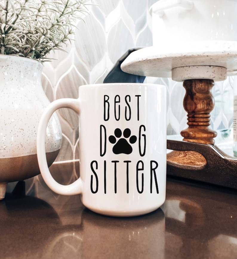 May include: White ceramic coffee mug with a black paw print and the text "Best Dog Sitter" in black.