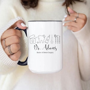 May include: White ceramic mug with a black rim and handle. The mug features a black and white illustration of dental tools, a tooth, and the text "Dr. Adams, Doctor of Dental Surgery."
