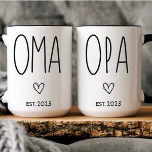 May include: Two white ceramic mugs with black rims and handles. The mugs have black text that reads "OMA EST. 2023" and "OPA EST. 2023" with a small heart symbol below the text.