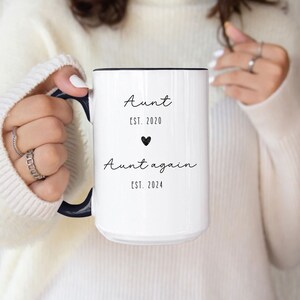 May include: White ceramic mug with a black rim and handle. The mug has black text that reads "Aunt Est. 2020" with a small heart symbol and "Aunt again Est. 2024".