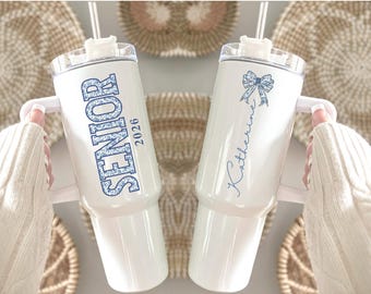 May include: Two white insulated tumblers with handles. One features the word "SENIOR" and the year "2026" in blue floral print. The other has a blue bow and the name "Katherine" in script. Both have straws.