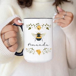 May include: White ceramic mug with a black handle. The mug features a bee with yellow and black stripes, a honeycomb, and floral designs. The name "Amanda" is printed on the mug.
