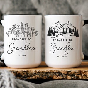 May include: Two white ceramic mugs with black handles and black rims. The mug on the left has a floral design and the text "Promoted to Grandma Est. 2024". The mug on the right has a mountain scene with the text "Promoted to Grandpa Est. 2024".