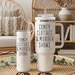 May include: Two white insulated tumblers with the text "COMFY CLOTHES & MURDER SHOWS" in black. One tumbler is 20oz, the other is 40oz. Both have a straw and a lid. The tumblers are on a woven tray.